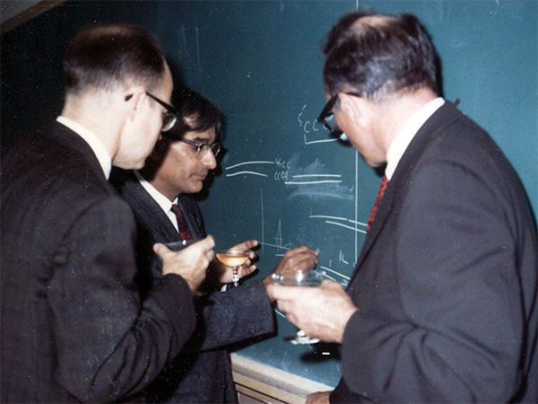 Khorana and two men writing on a chalkboard, champagne in hand