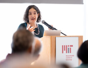 Laura Schulz stands at a podium delivering her lecture to the audience, whose blurry heads appear in the foreground.